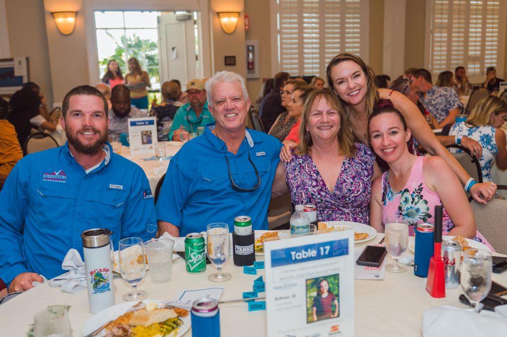 A group of five people sit and smile at a dining table labeled "Table 17" during an indoor event, with food and drinks in front of them.