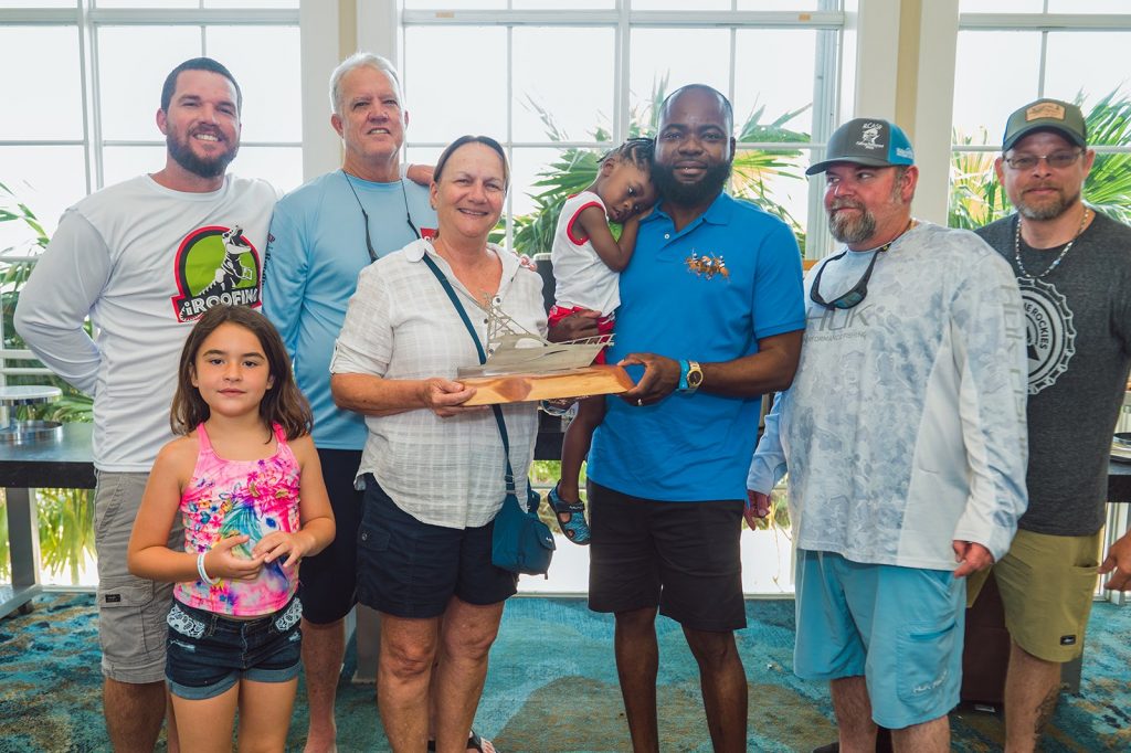 A group of seven adults and two children pose indoors with a wooden award, standing in front of large windows and tropical plants.