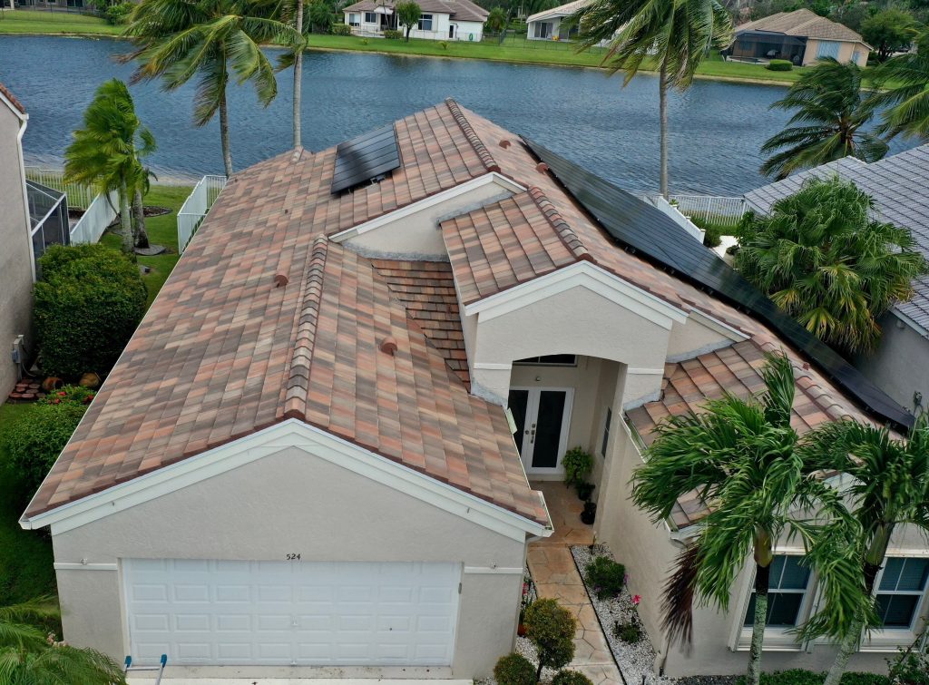 Aerial view of a beige house with brown tiled roof and solar panels, surrounded by palm trees, with a lake and neighboring houses in the background.