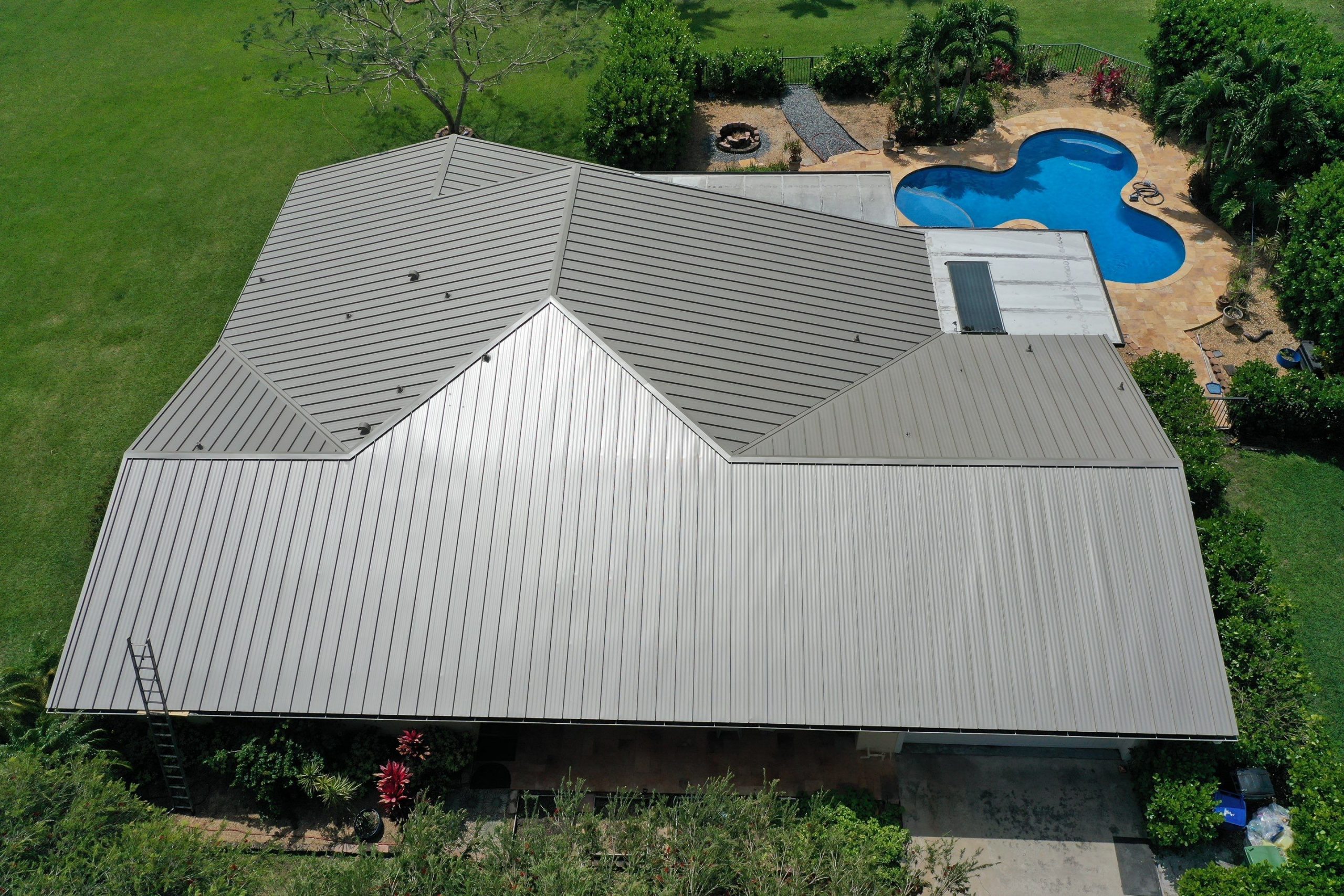 Aerial view of a house with a metal roof, surrounded by green lawn and trees, and a uniquely shaped blue swimming pool in the backyard.
