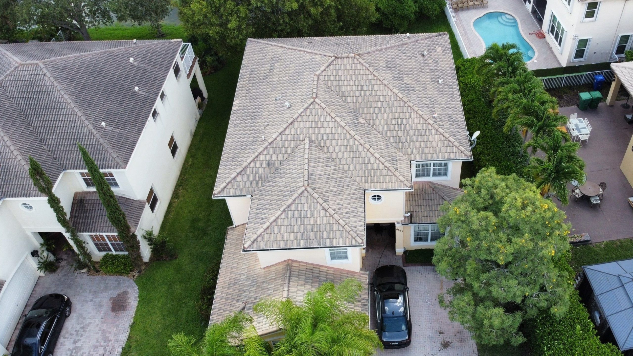 Aerial view of a two-story house with a tiled roof, driveway with a black car, green lawn, and neighboring homes; a swimming pool is visible in the upper right corner.