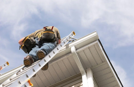 A worker wearing a tool belt stands on a ladder, inspecting or repairing the roof and gutter of a house under a partly cloudy sky.