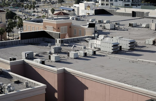 Rooftop view of a commercial building with multiple HVAC units and vents, surrounded by other buildings and a few visible palm trees in the background.
