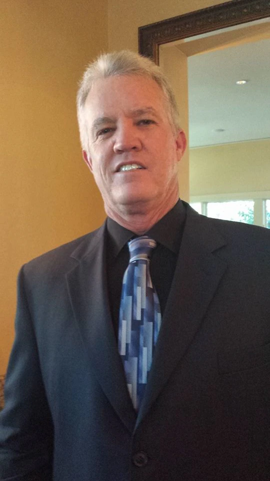A man with short gray hair wearing a dark suit, black shirt, and blue patterned tie stands indoors in front of a yellow wall and mirror.