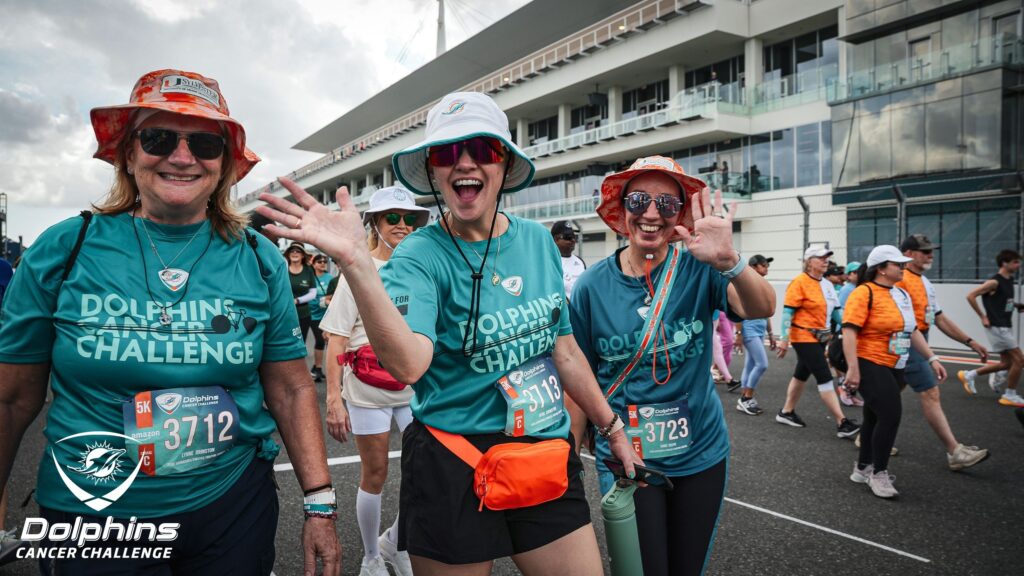 Three women wearing "Dolphins Cancer Challenge" shirts and hats smile and wave while walking in an outdoor event; other participants walk in the background.