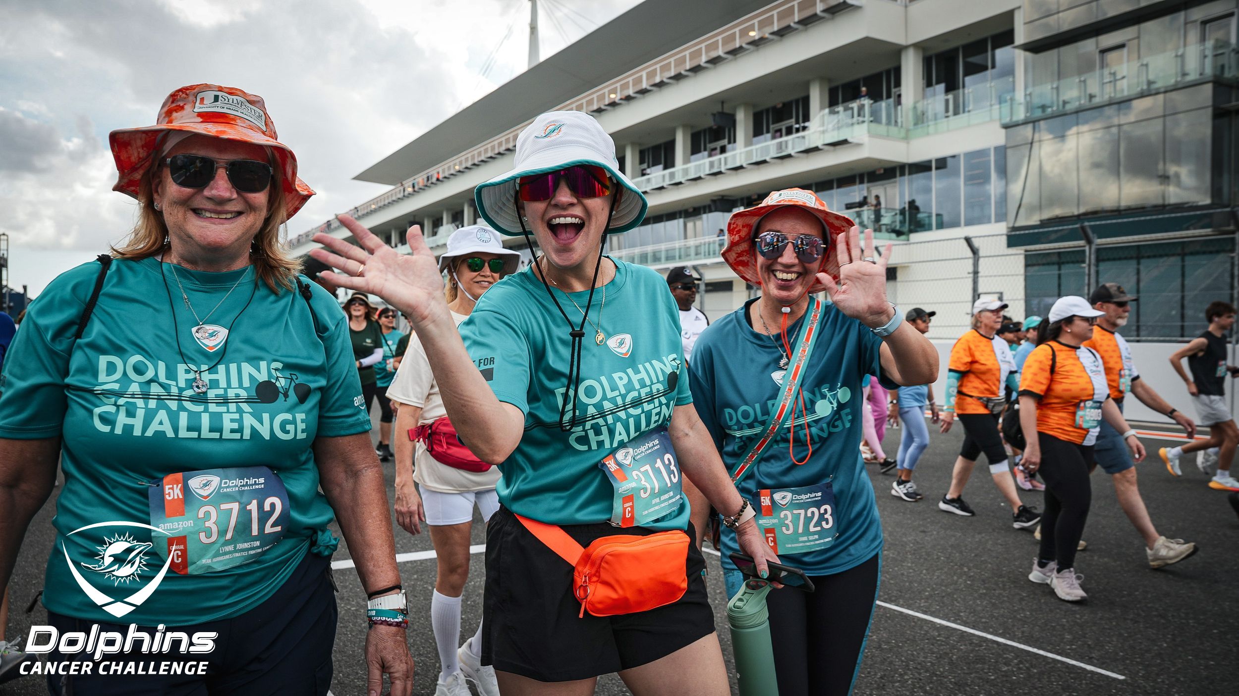 Three women wearing "Dolphins Cancer Challenge" shirts and hats smile and wave while walking in an outdoor event; other participants walk in the background.