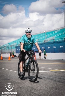 A man wearing a helmet and teal Dolphins Challenge t-shirt rides a bicycle on a racetrack. Orange cones and a stadium are in the background.