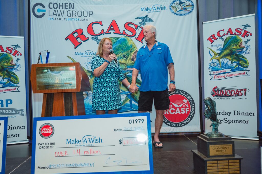 A woman and man stand on stage holding hands, speaking into a microphone, with large check and fishing tournament banners in the background.