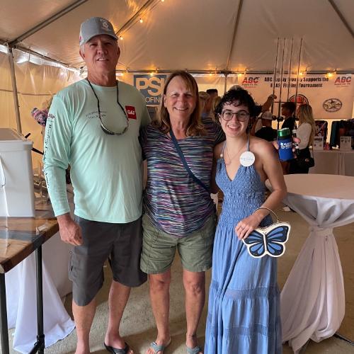 Three people stand smiling together inside a tent at an event. The background features informational tables, banners, and event displays.