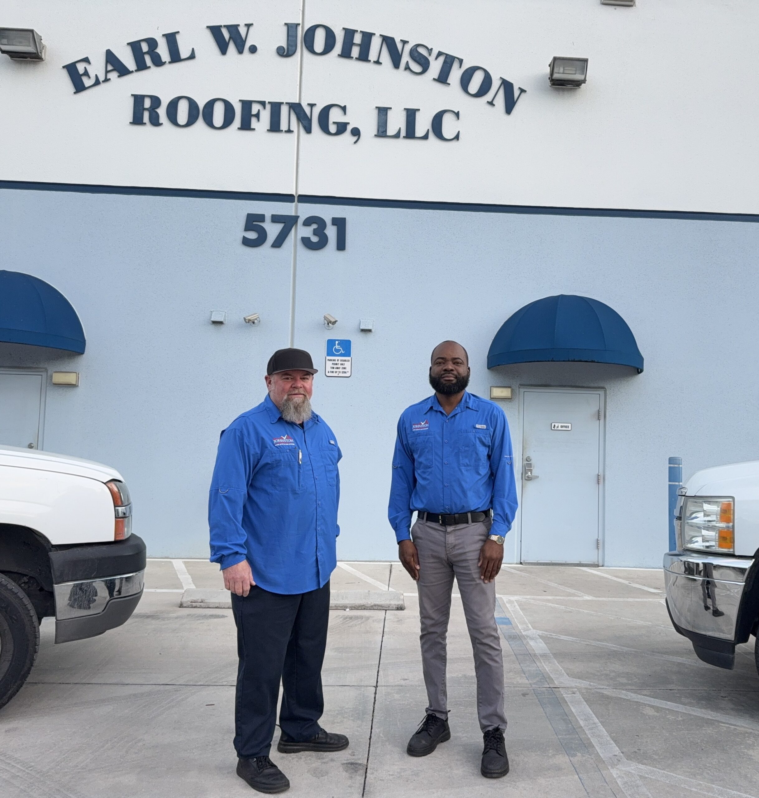 Two men in blue shirts stand in front of Earl W. Johnston Roofing, LLC building, next to two white trucks in a parking lot.
