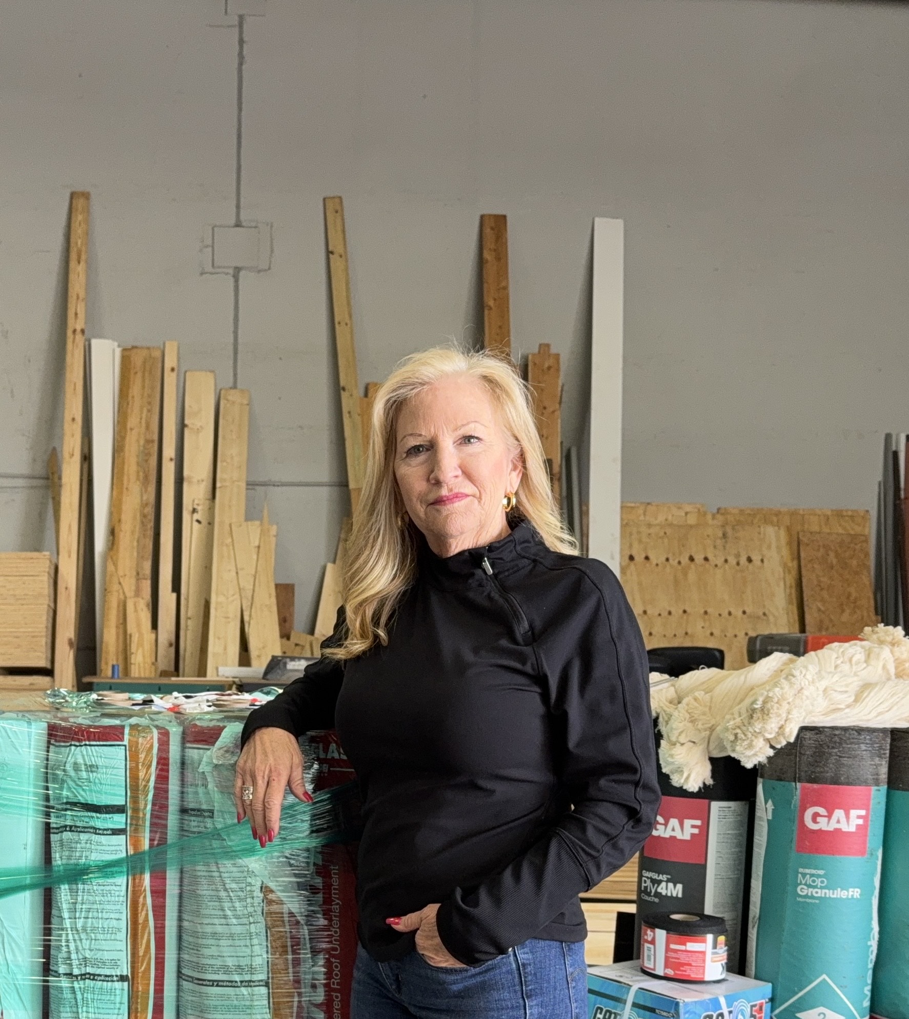 A woman in a black jacket stands in a warehouse, surrounded by stacks of construction materials, including wood planks and roofing supplies.