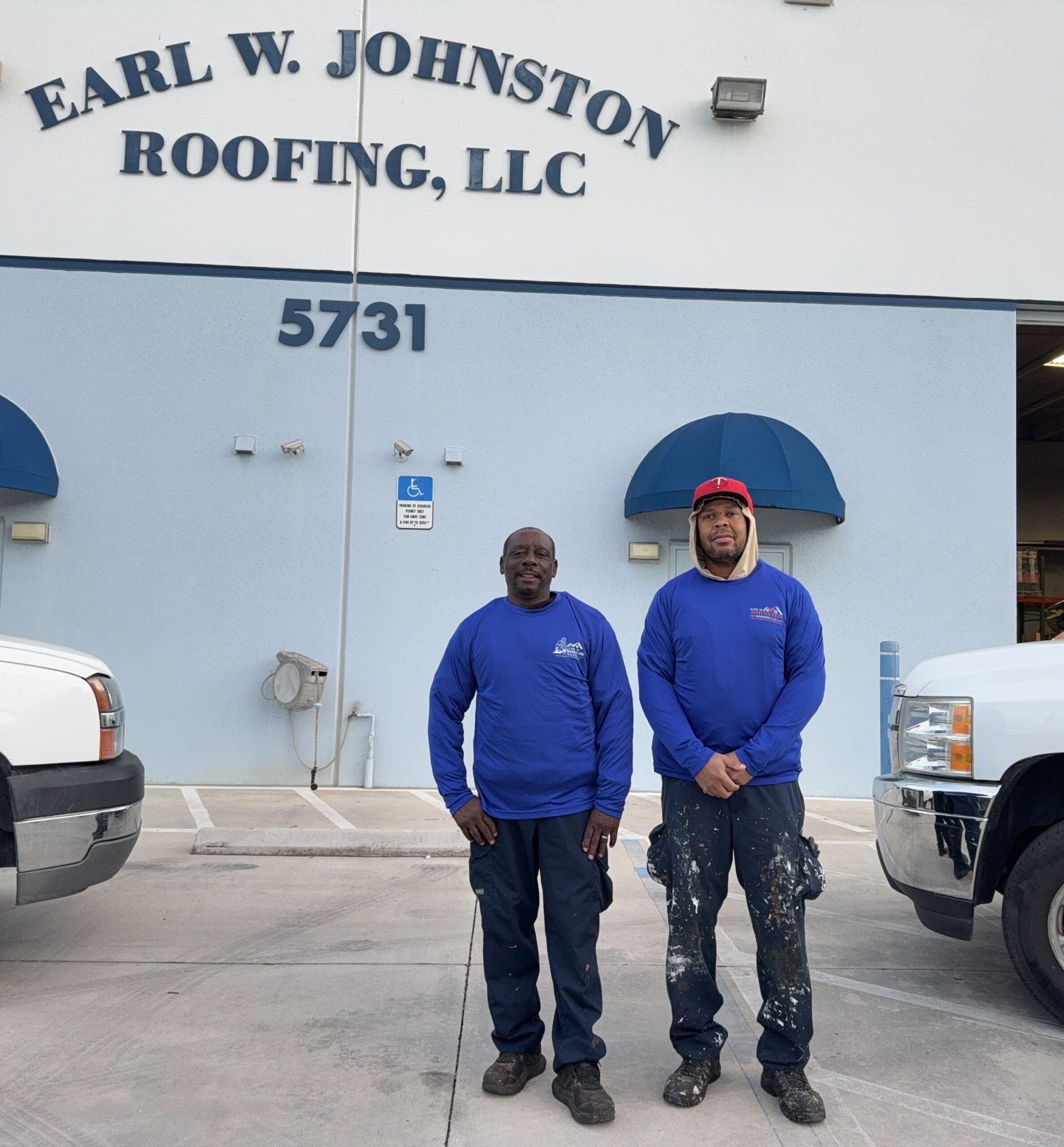 Two men in blue work shirts stand in front of a building labeled "Earl W. Johnston Roofing, LLC" with the street number 5731, between two white trucks.