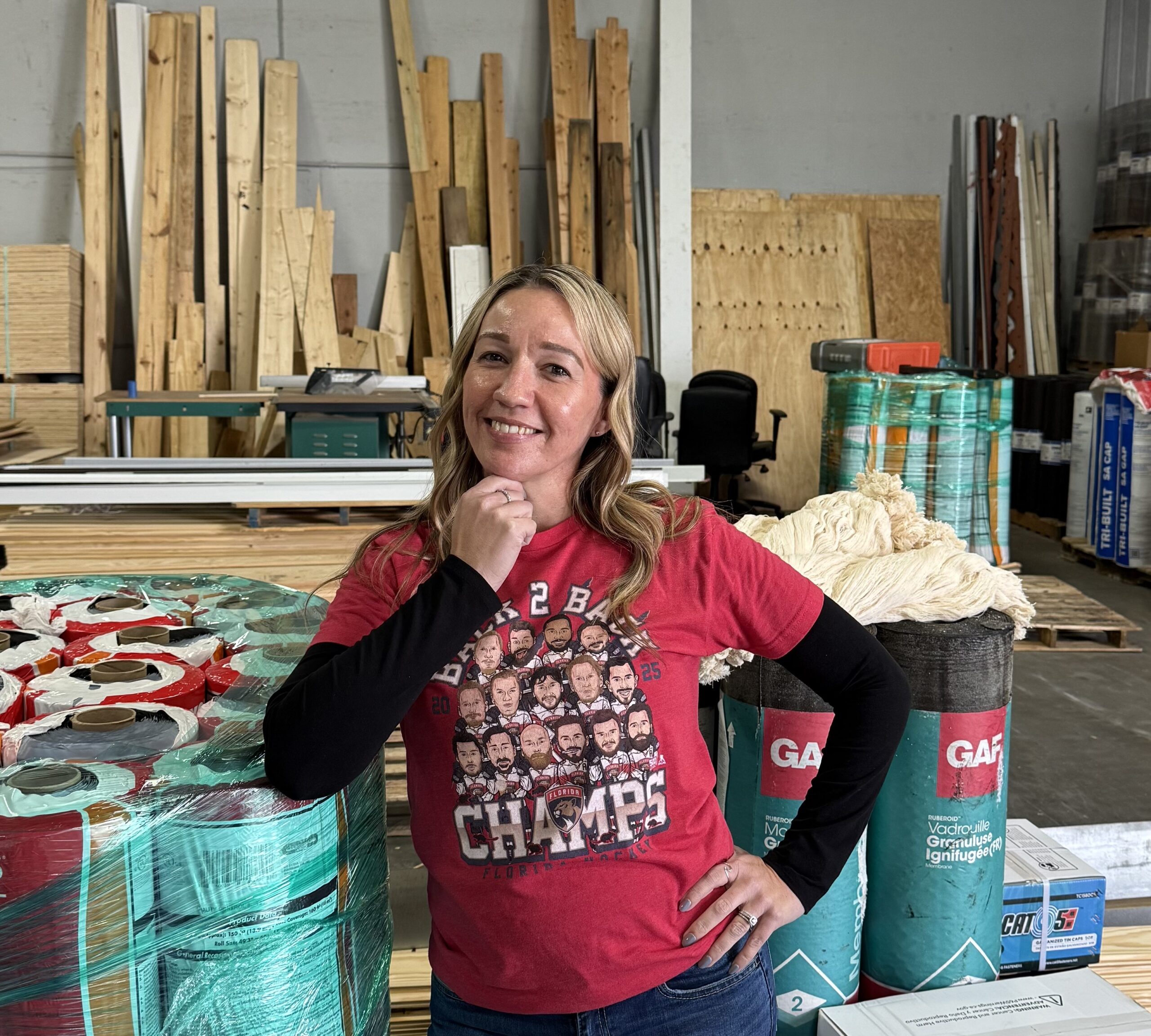 A woman in a red T-shirt stands in a warehouse with stacks of construction materials and wood in the background.