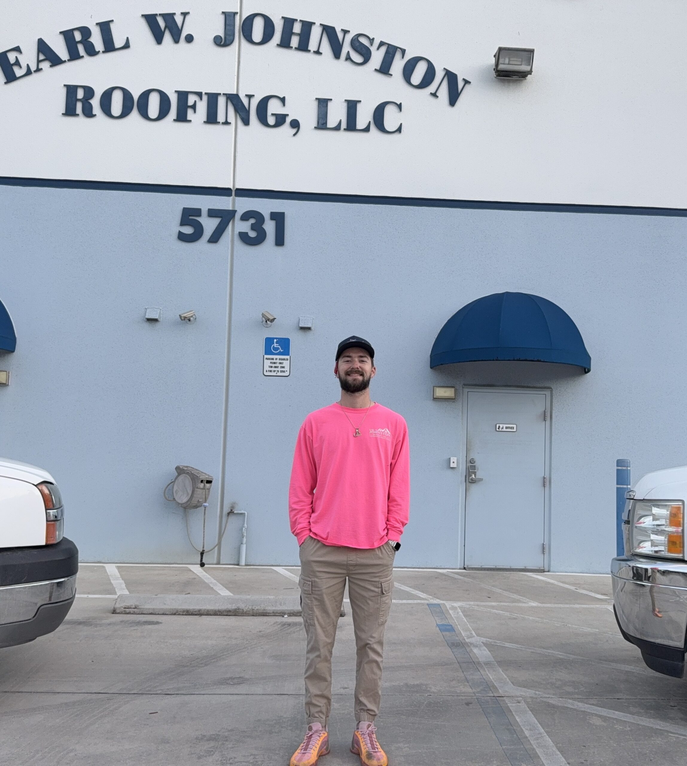 A man in a bright pink long-sleeve shirt stands in front of a building labeled "Earl W. Johnston Roofing, LLC" and the number 5731, with two white trucks parked nearby.