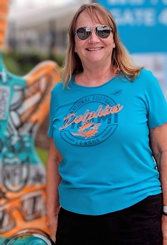 Smiling woman in sunglasses and a Miami Dolphins t-shirt stands outdoors with a colorful painted sculpture in the background.
