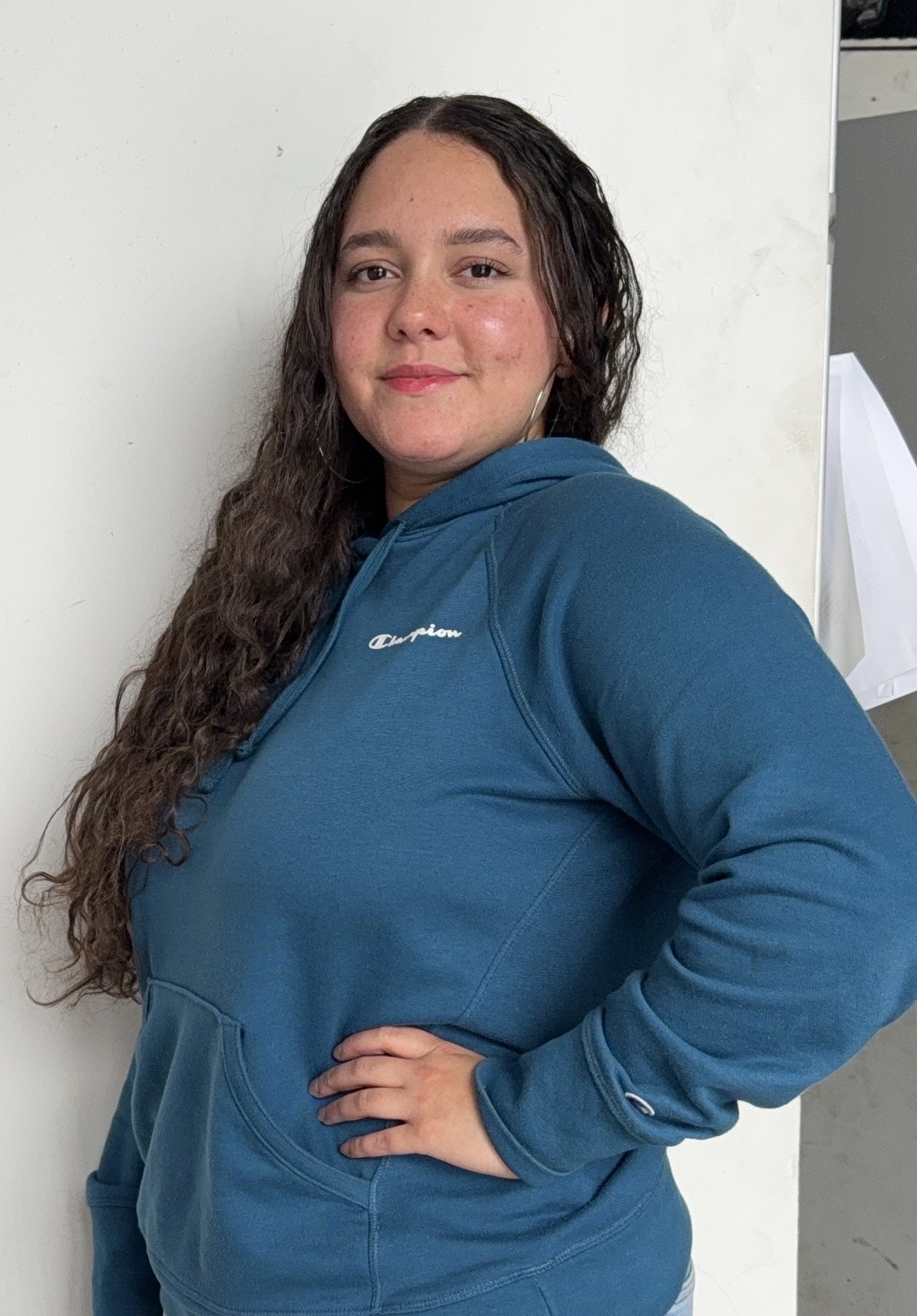 A woman with long curly hair wearing a teal Champion hoodie stands against a white wall, looking at the camera with a neutral expression.