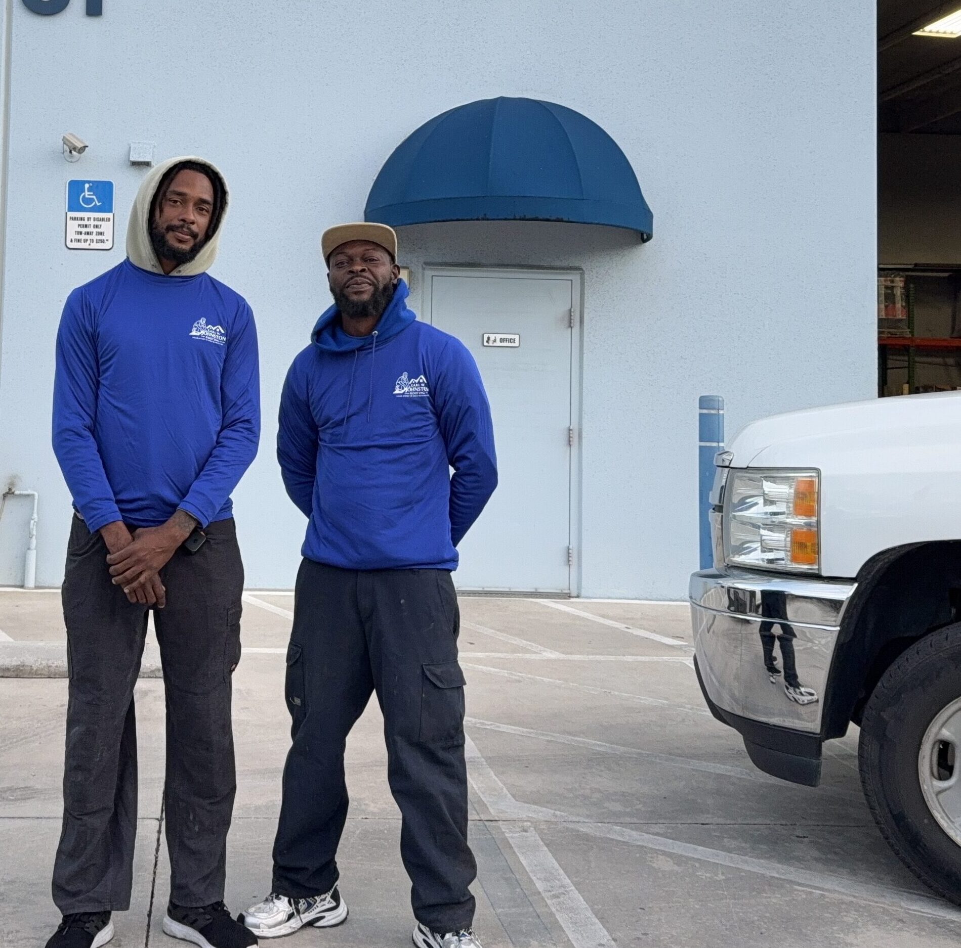 Two men in blue work uniforms stand outside Earl W. Johnston Roofing, LLC, beside two white trucks, in front of building number 5731.